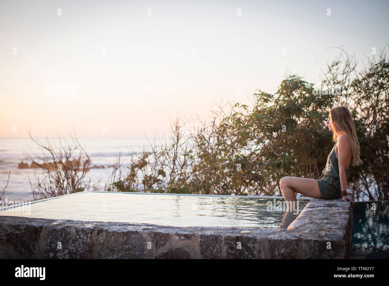 Side view of woman sitting on retaining wall at artificial pond against sea and clear sky Stock Photo