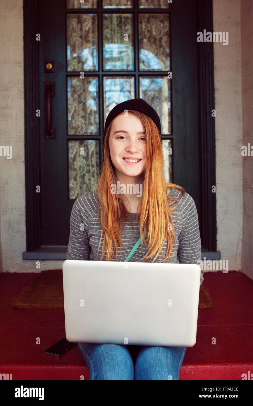 Portrait of happy teenage girl sitting with laptop on steps outside house Stock Photo