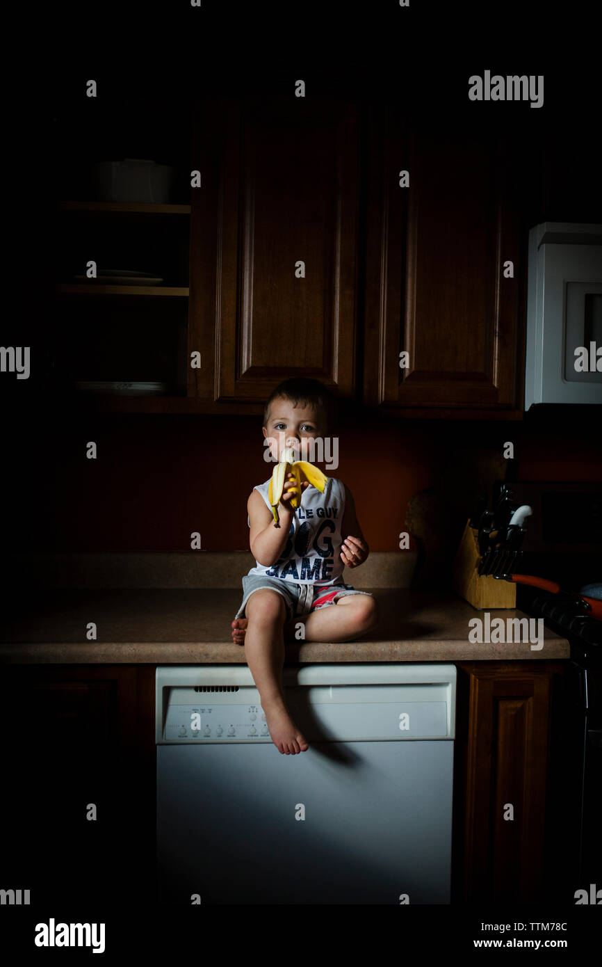Portrait of boy eating banana while sitting on kitchen counter at home Stock Photo