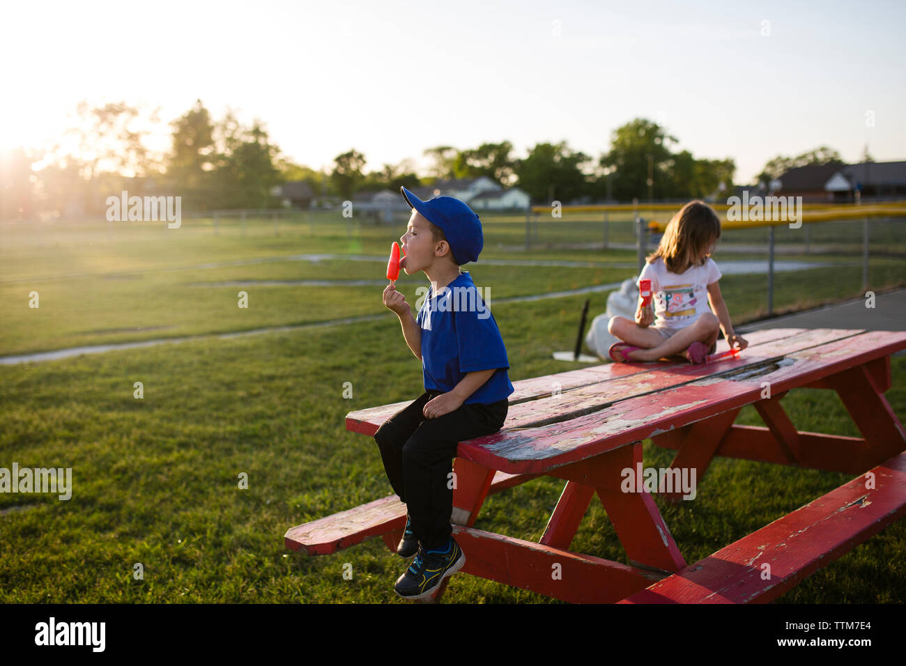Siblings eating flavored ice while sitting on picnic bench at park Stock Photo