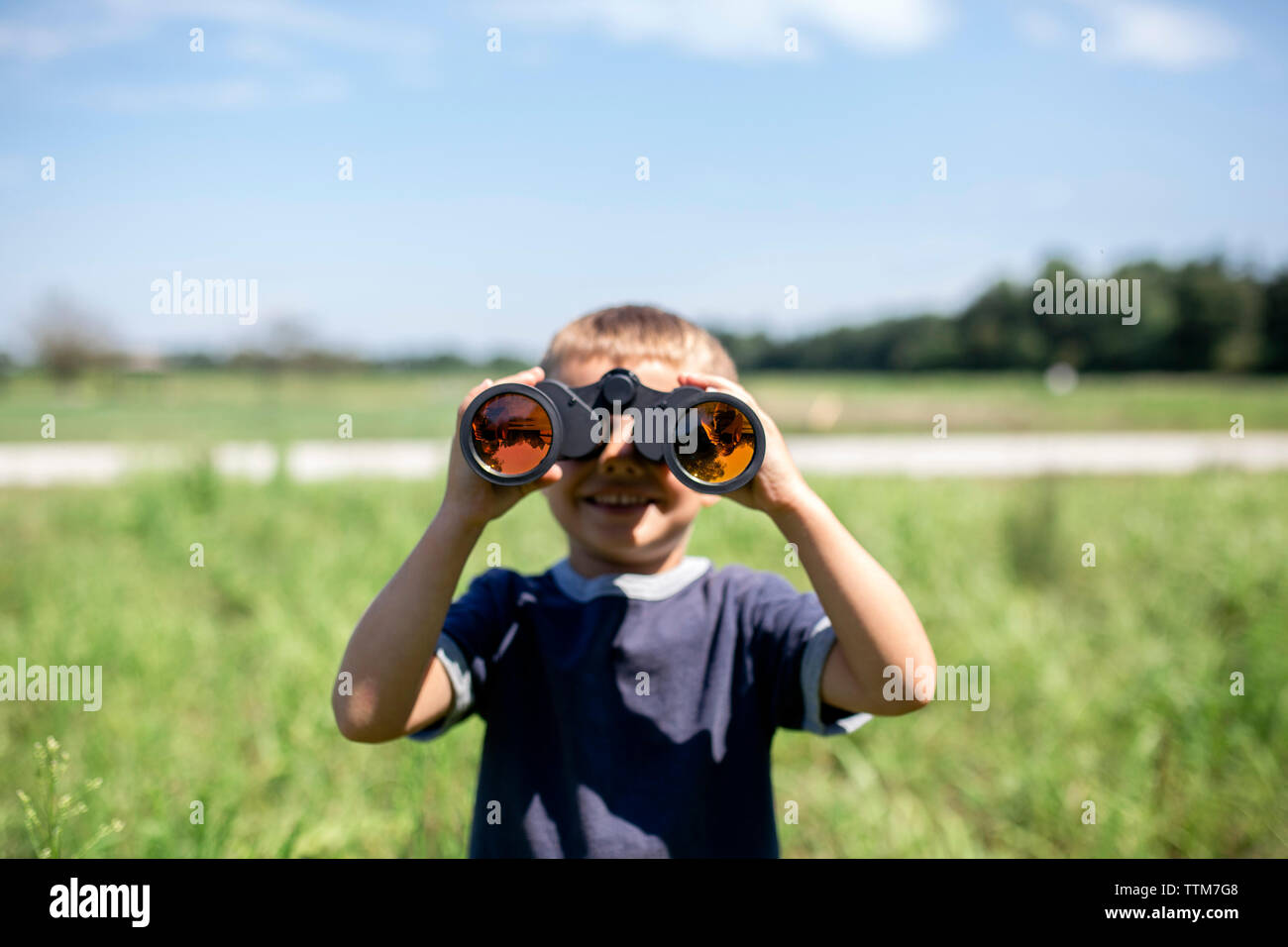 Playful boy looking through binoculars while standing on field during sunny day Stock Photo