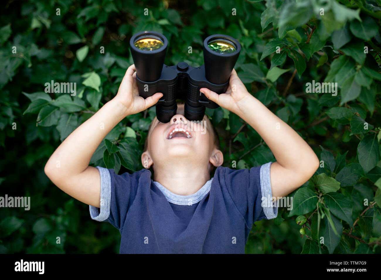 Playful boy looking through binoculars while standing against plants Stock Photo