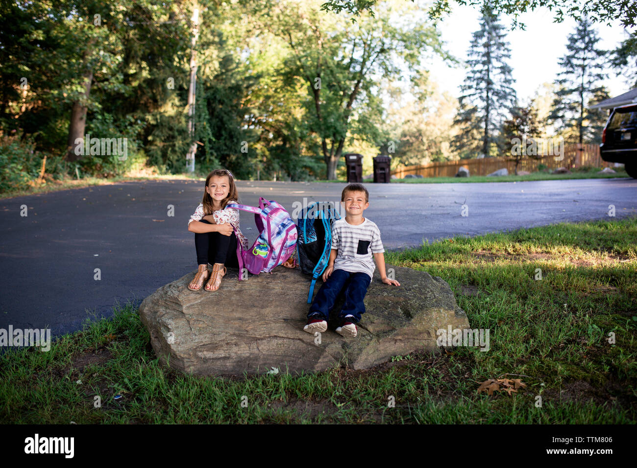 Brother and sister siblings sitting waiting on rock for school bus Stock Photo