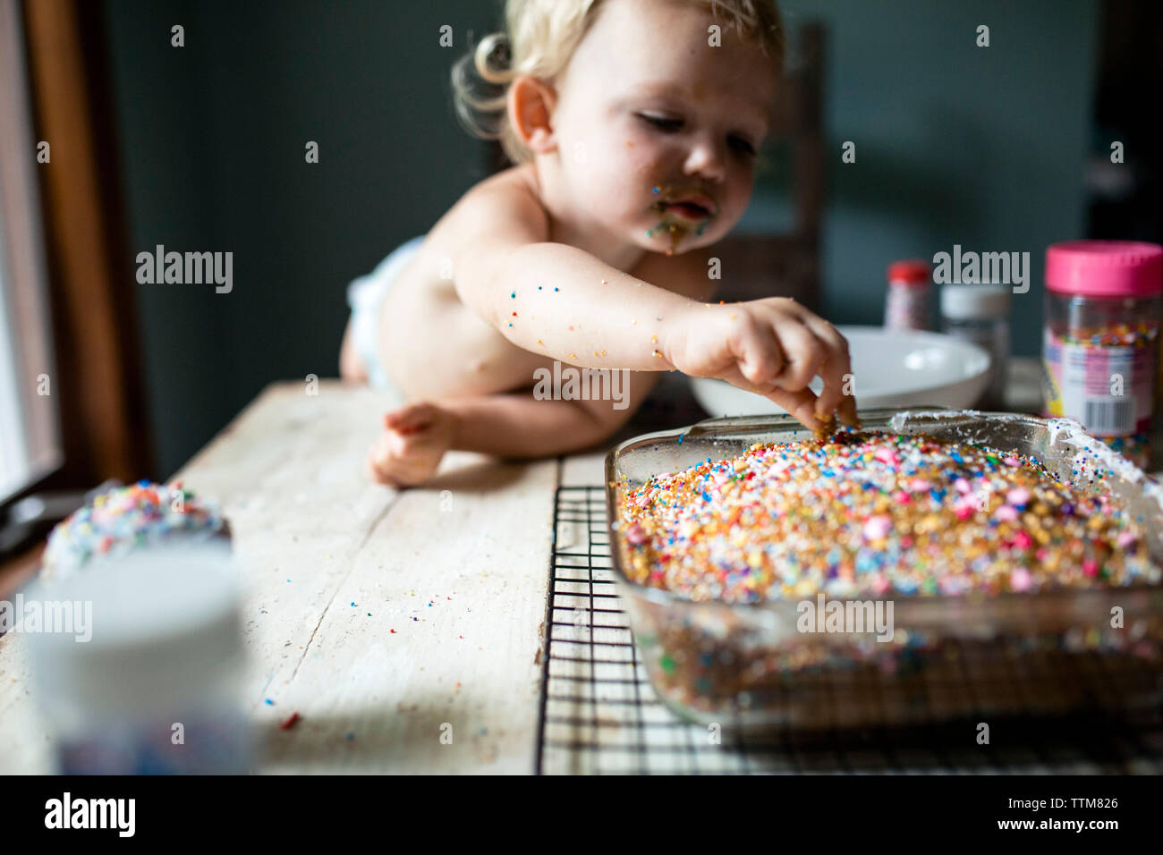 Toddler girl sneaking colorful sprinkles off of freshly baked cake Stock Photo