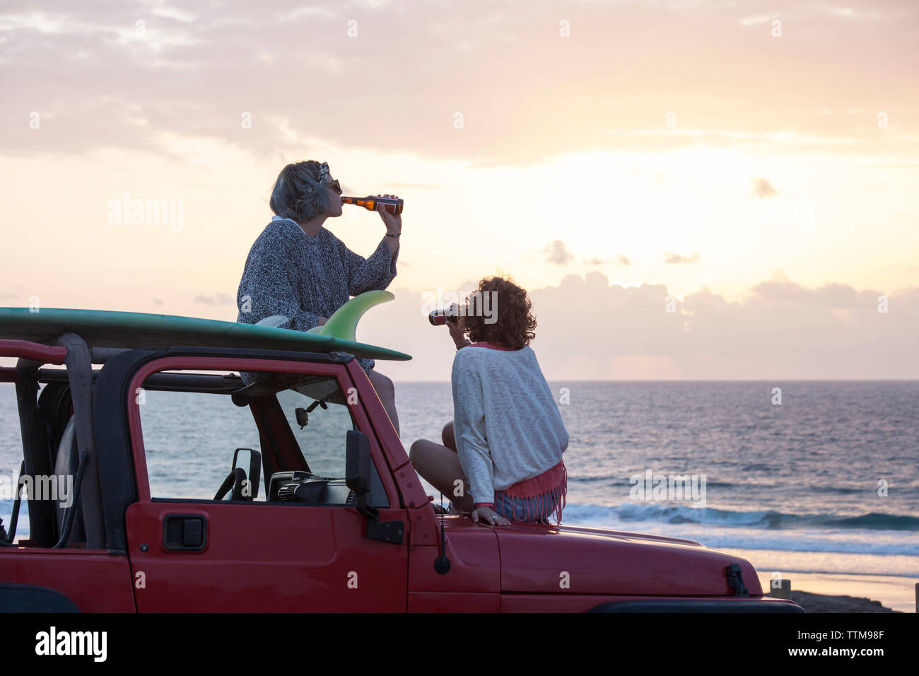 Two surfer girls with sunglasses on their 4x4 car drinking beer Stock Photo