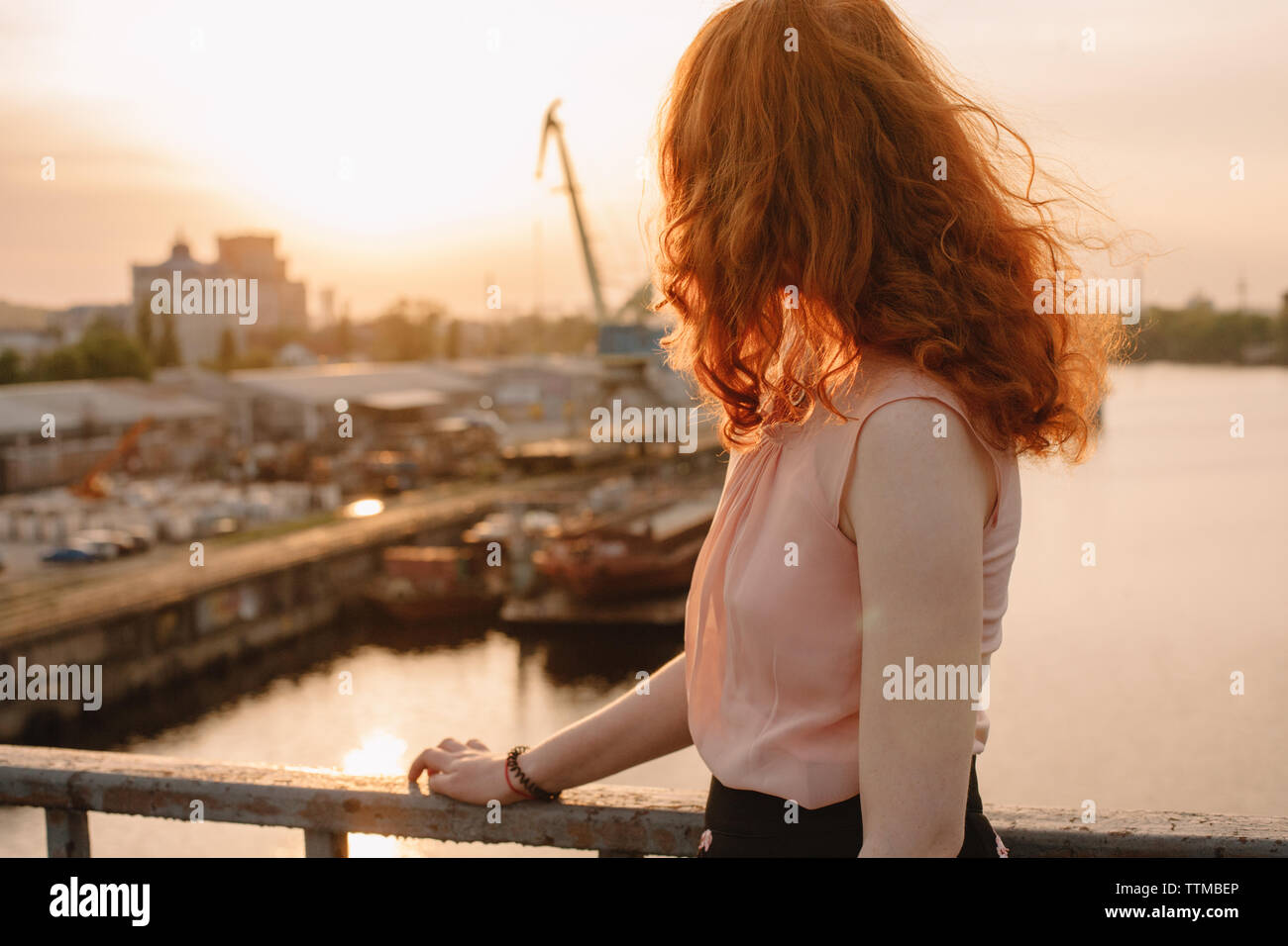 Young woman with red hair standing by railing on bridge at sunset Stock Photo