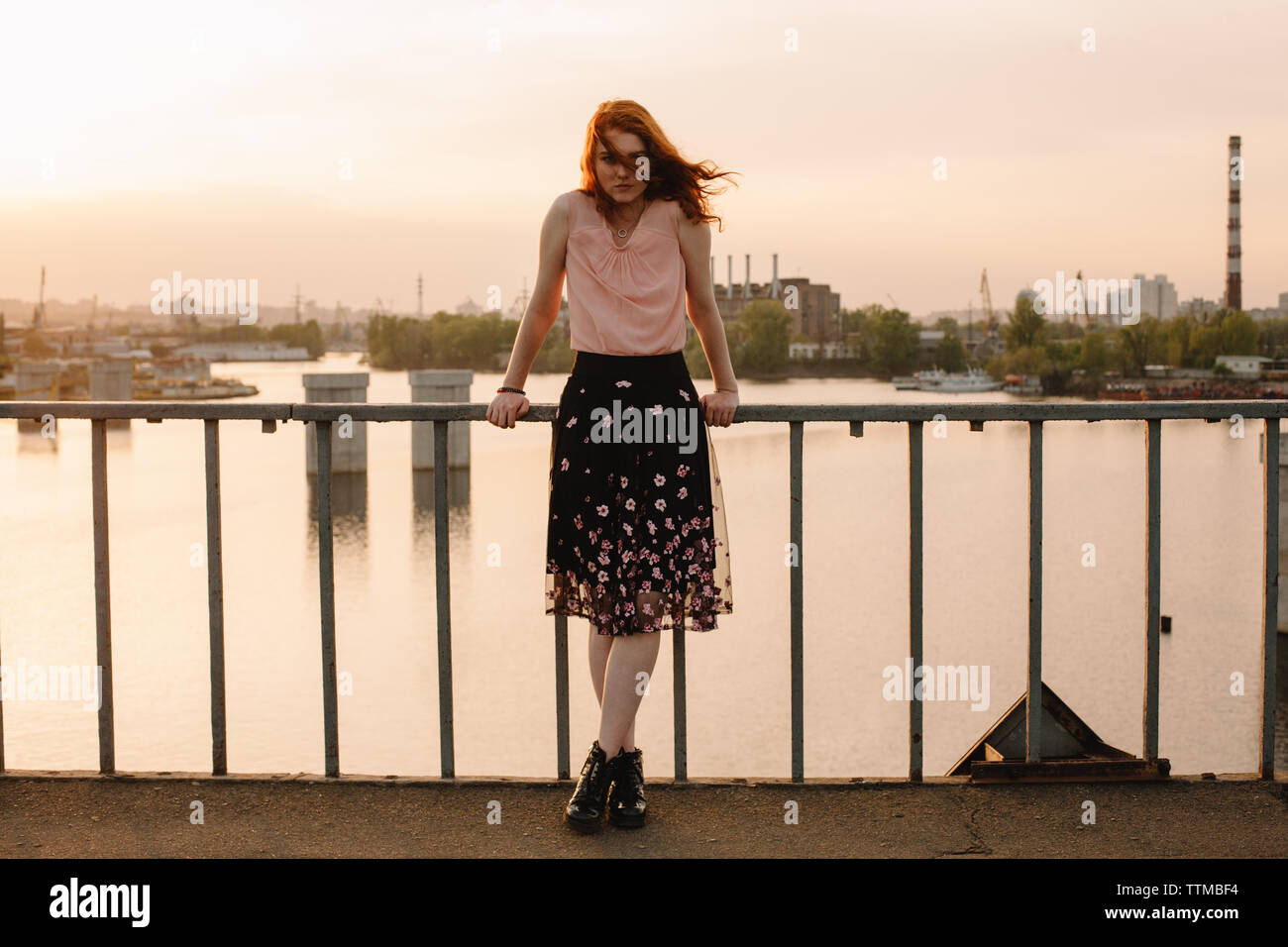 Young woman with red hair standing by railing on bridge against river ...