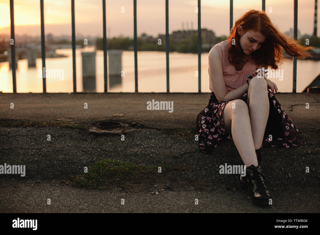 Young redhead woman using smart phone while sitting on bridge in city Stock Photo