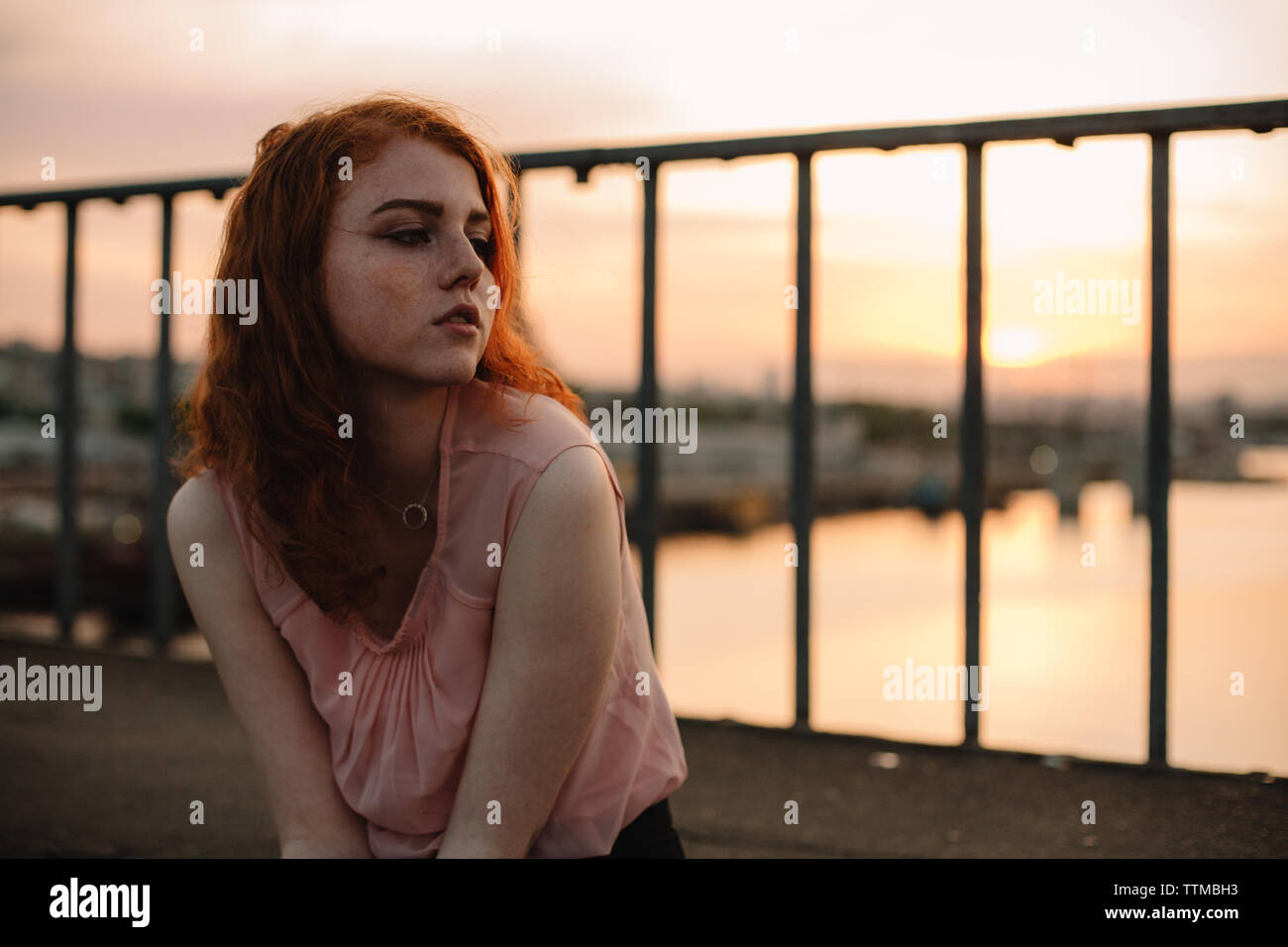 Portrait of young red head woman sitting on bridge at sunset Stock Photo