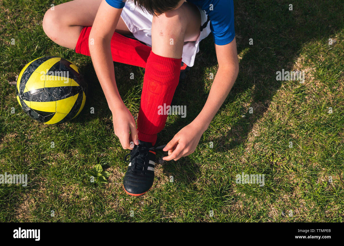 High angle view of boy tying shoelace while sitting by soccer ball on grassy field Stock Photo