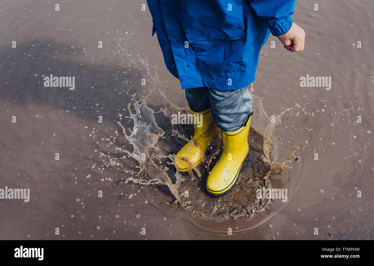 Low section of playful boy wearing yellow rubber boots while splashing puddle Stock Photo