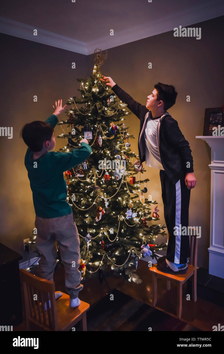 Two Boys Standing On Chairs To Decorate The Top Of A Christmas