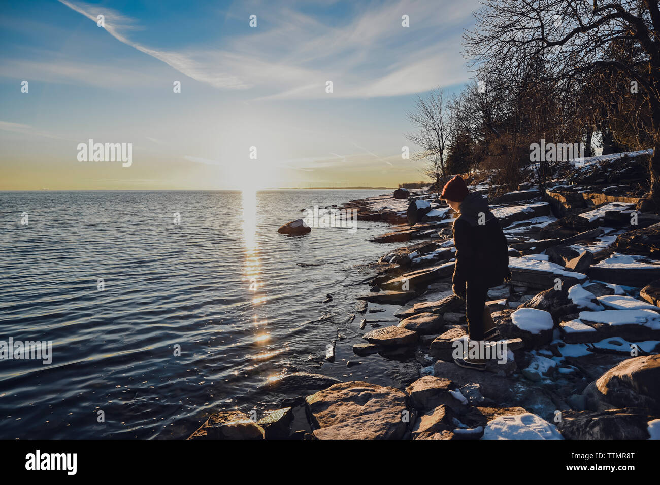 Boy standing alone on snowy rocks on lakeshore at sunset in winter. Stock Photo