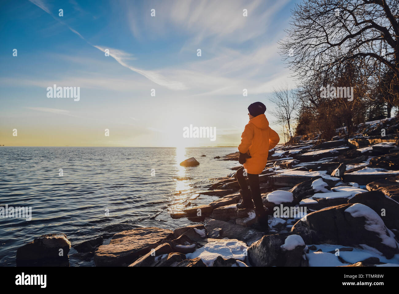 Boy standing alone on shoreline of lake at sunset on a winter evening. Stock Photo