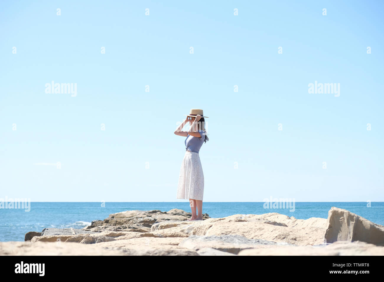 Young woman wearing hat while standing on rocks at beach against clear sky during sunny day Stock Photo