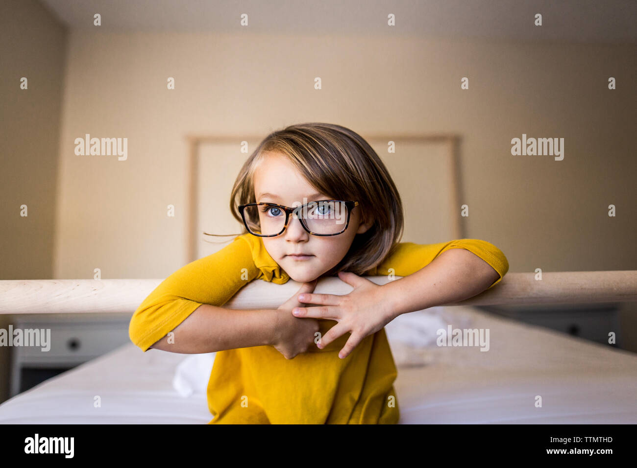 Young girl with glasses and yellow dress sitting on bed Stock Photo