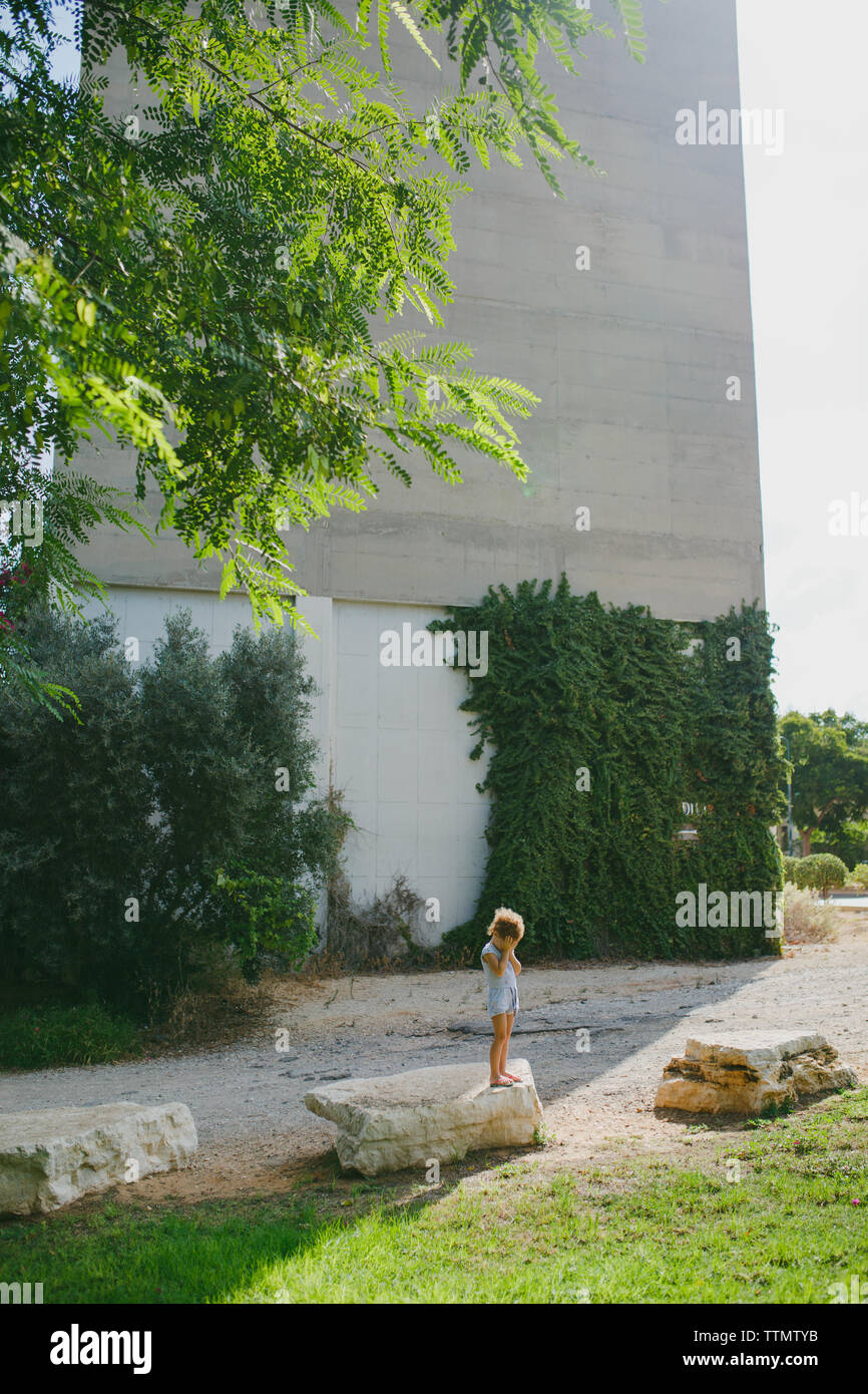A young girl covering her face while standing on a stone outside Stock Photo