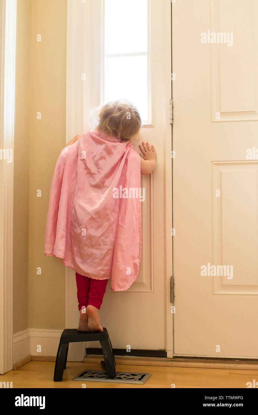 Rear View Of Girl Wearing Cape Standing On Stool By Door At Home Stock