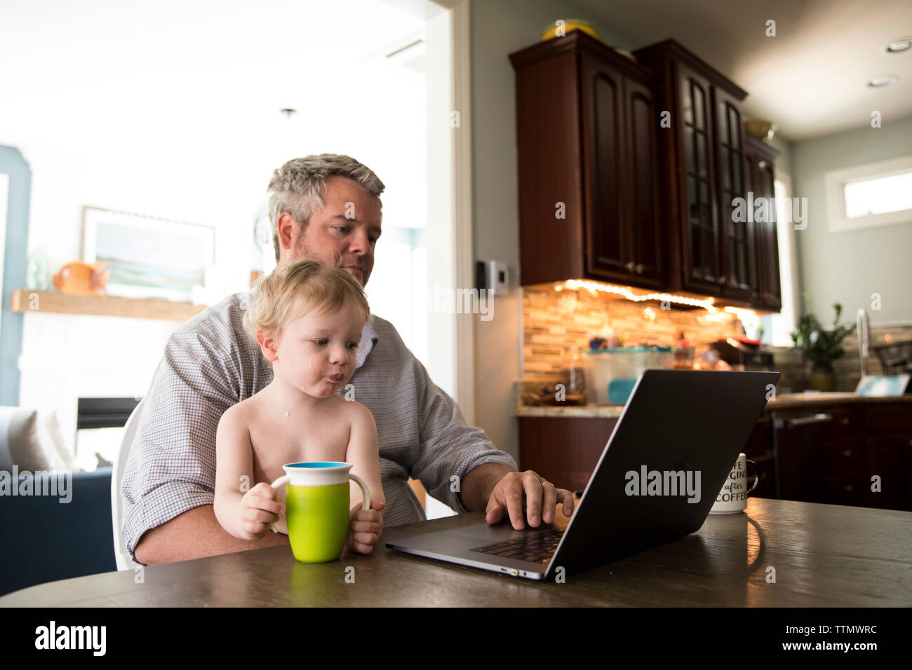 Work at Home Dad Sitting at Computer in Kitchen, Holding Toddler Son Stock Photo