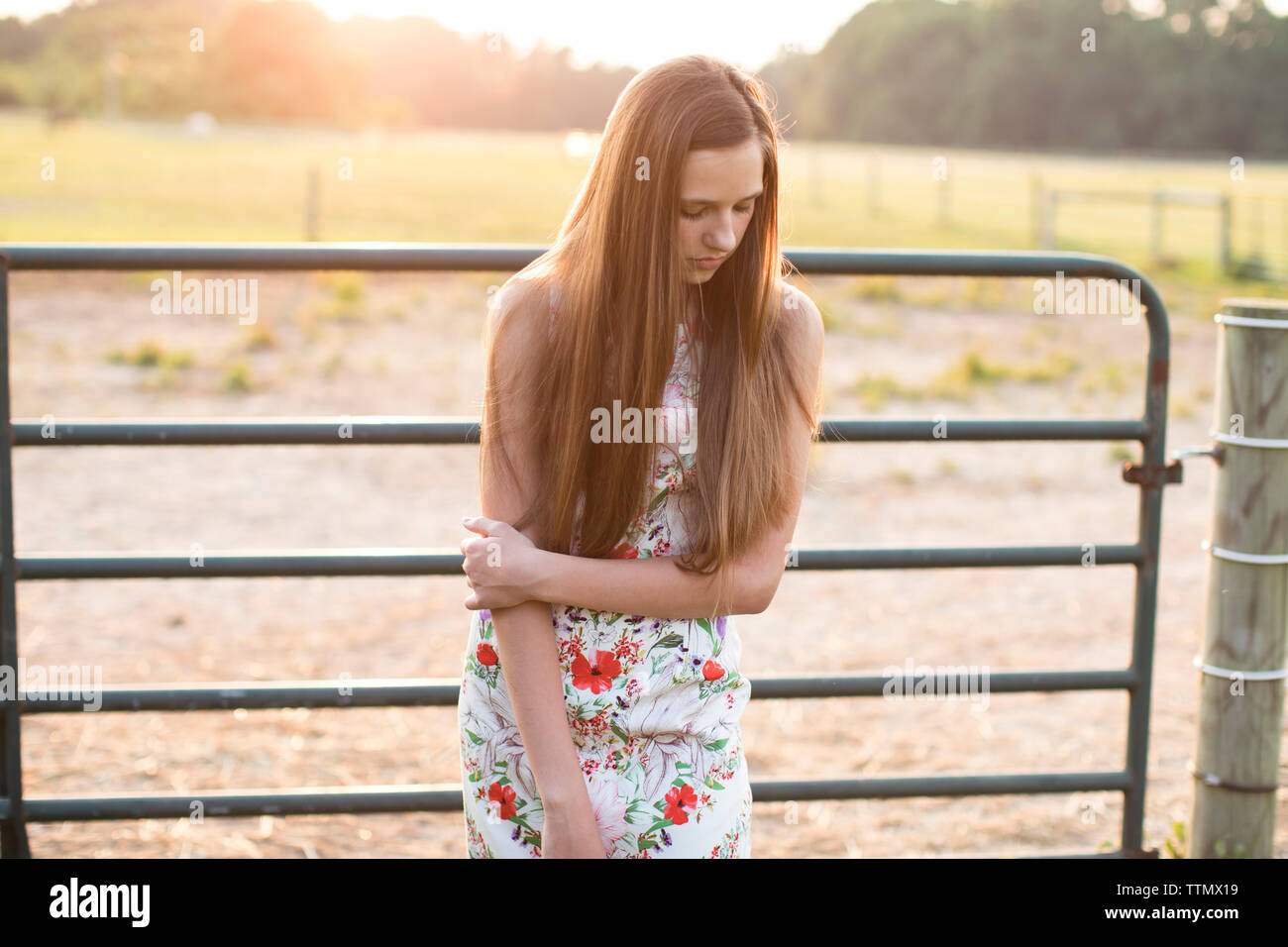 Girl Standing in Front of a Horse Farm Fence at Sunset, Looking Down Stock Photo