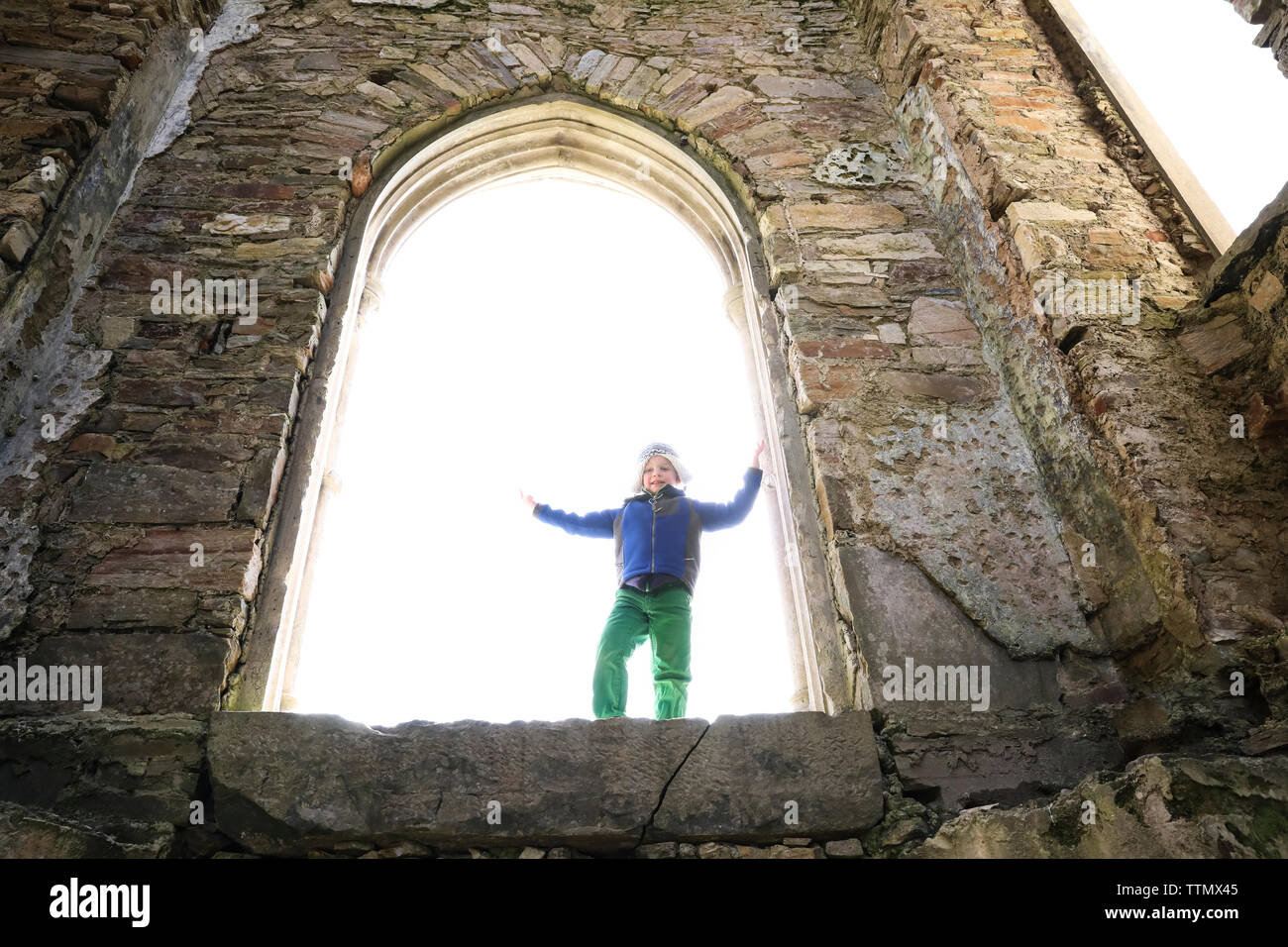 Backlit Young Boy Stands in Large Window in Stone Castle Stock Photo