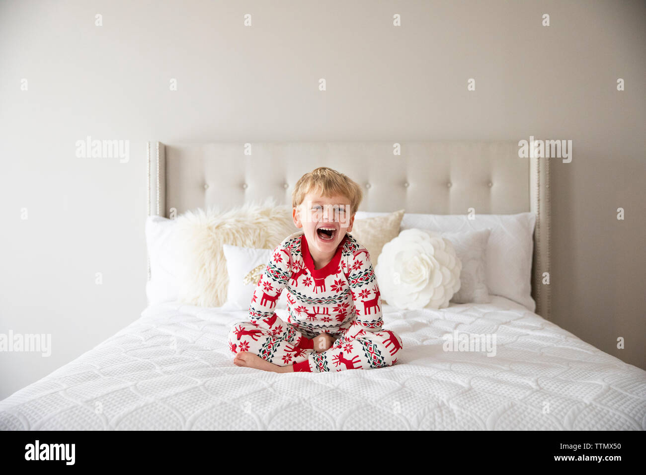 Cute Blonde Boy Wearing Holiday Pajamas Laughs While Sitting on Bed Stock Photo