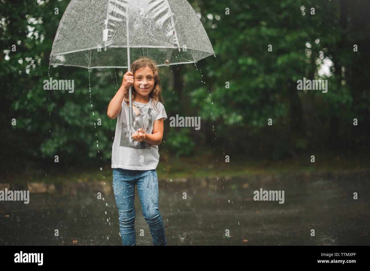 Portrait of girl carrying umbrella while walking on road against trees during rainfall Stock Photo