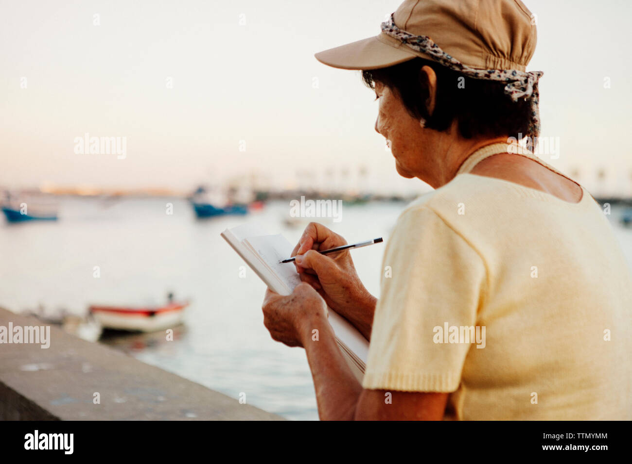 Senior woman drawing on book while sitting at retaining wall by sea Stock Photo