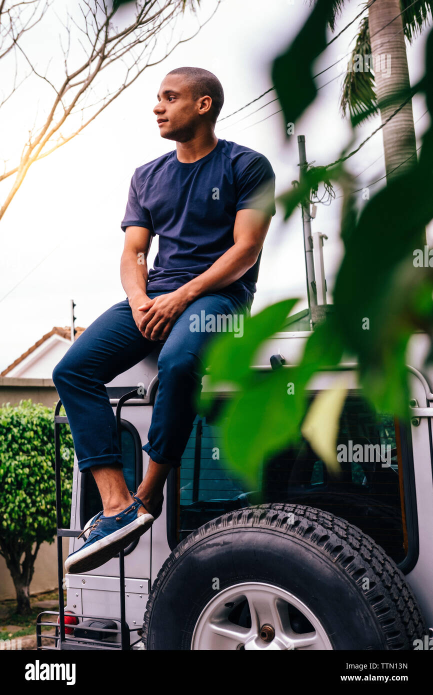 Full length of young man looking away while sitting on SUV's rooftop Stock Photo