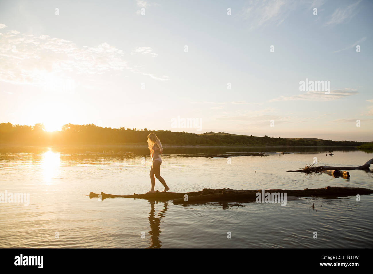 Side view of young woman in bikini standing at riverbank against sky during sunset Stock Photo