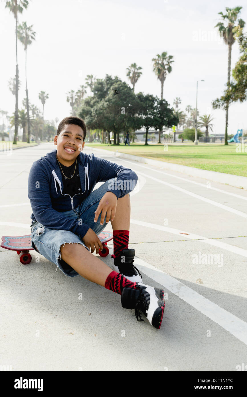 Full length portrait of smiling teenage boy sitting on skateboard at park Stock Photo