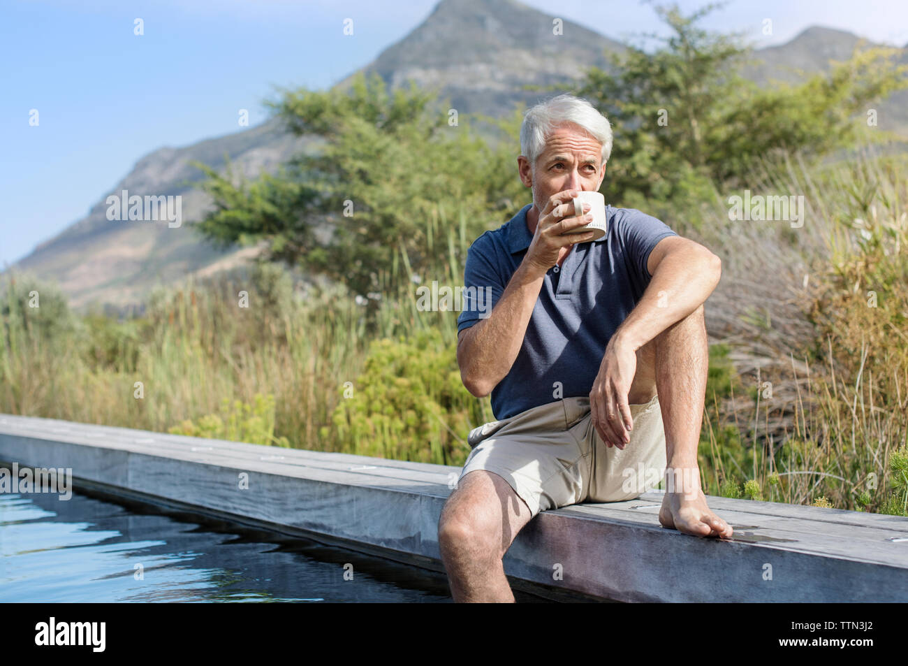 Mature man drinking coffee while sitting by swimming pool against mountain Stock Photo