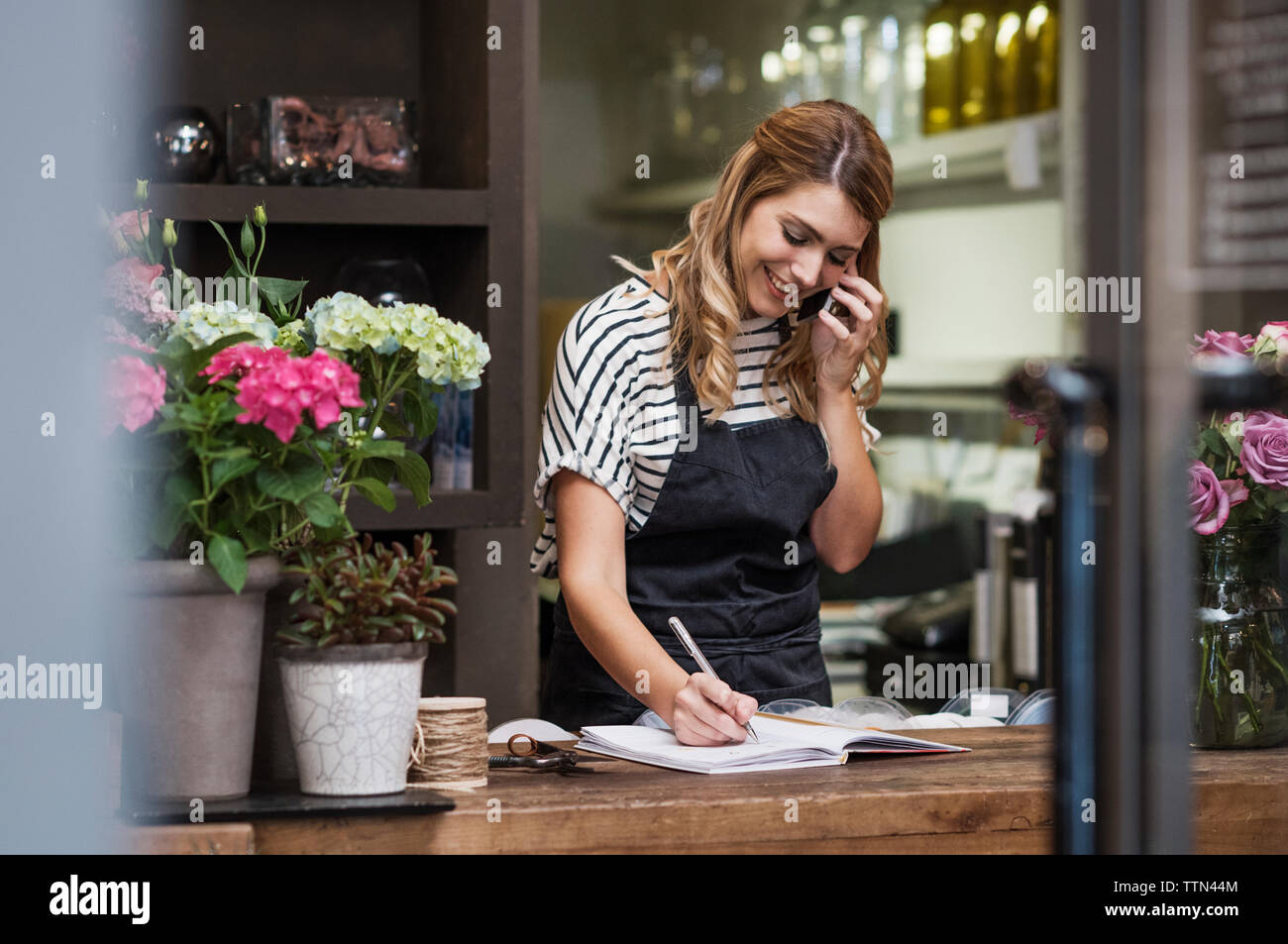 Florist writing in note pad while using mobile phone Stock Photo