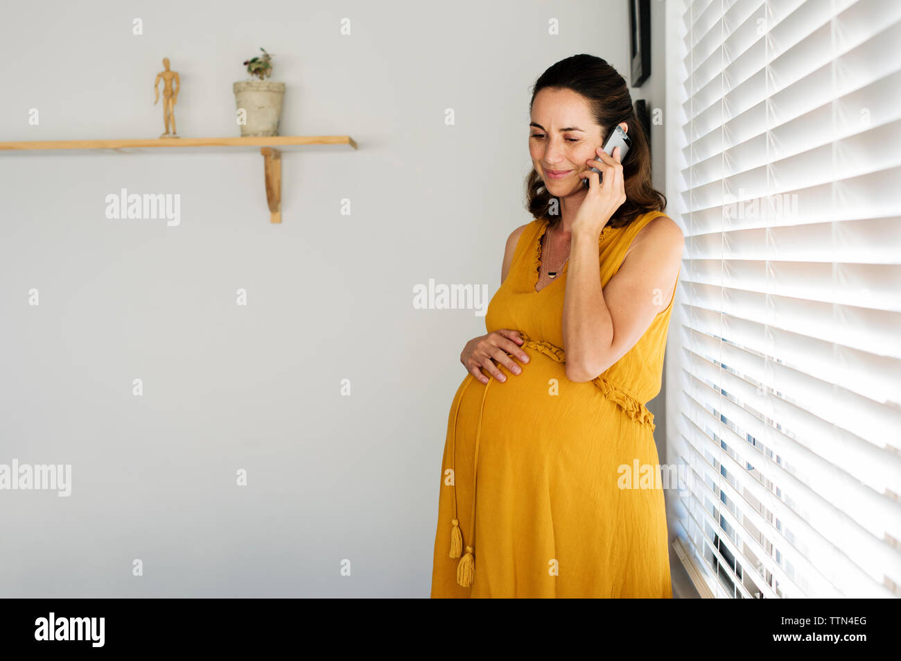 Pregnant woman talking on smart phone while standing by window at home Stock Photo