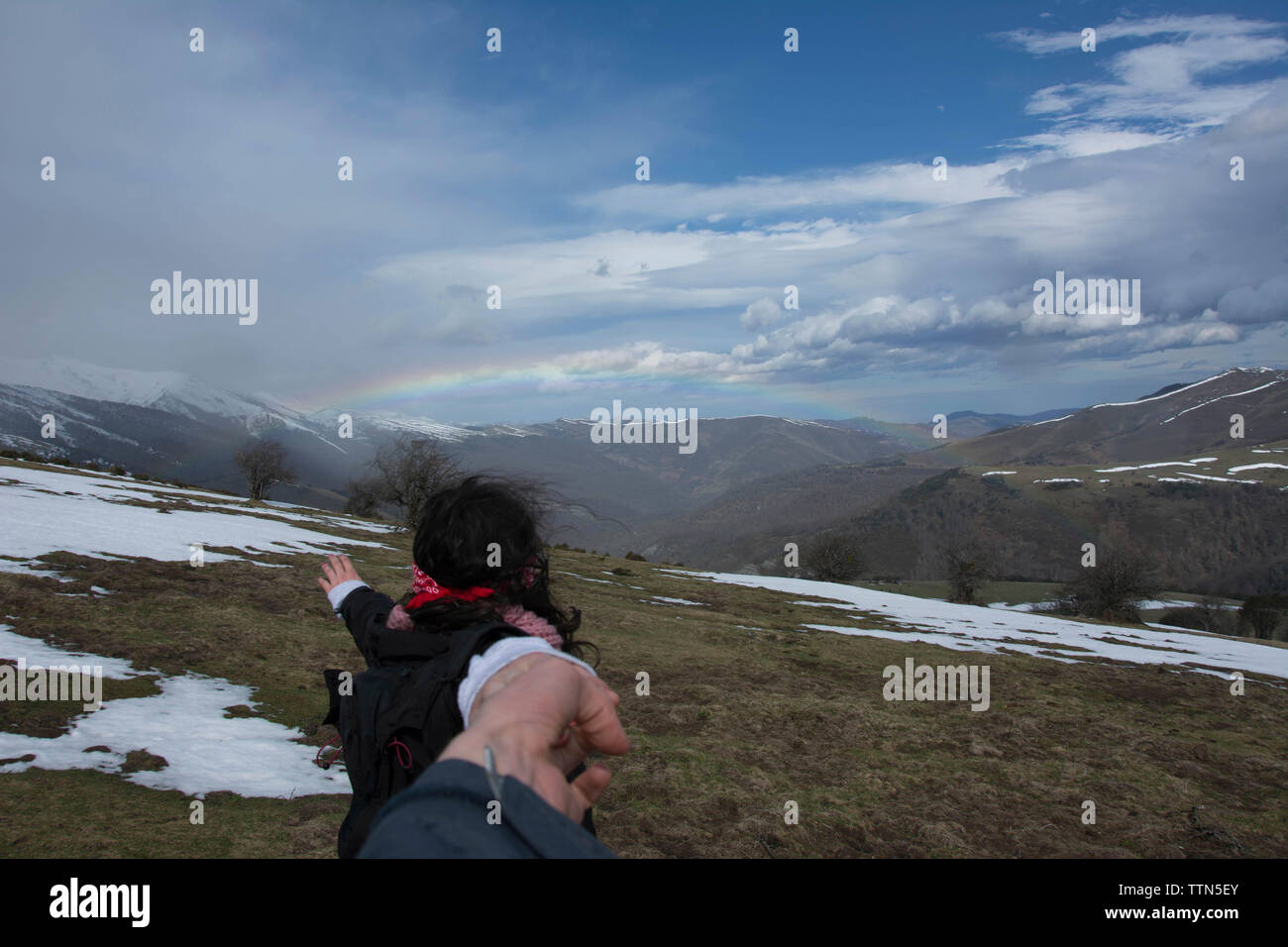 Cropped image of friend holding woman's hand against mountains and rainbow during winter Stock Photo