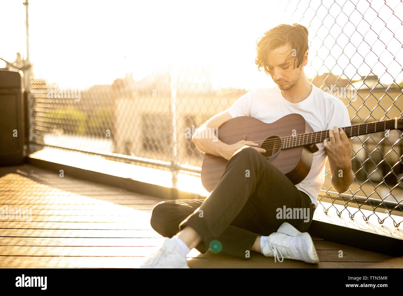 Man playing guitar while sitting by chainlink fence against sky during sunset Stock Photo