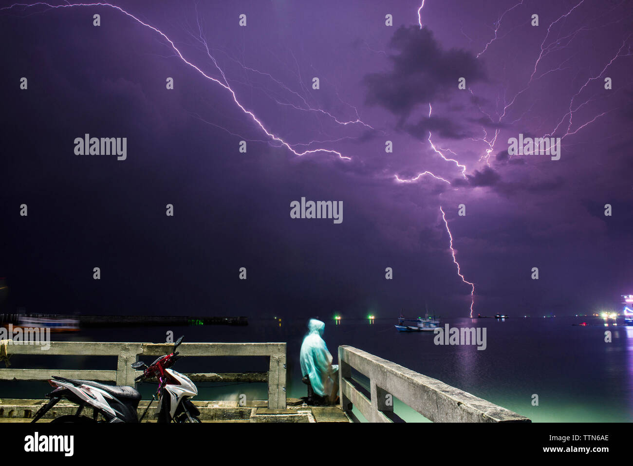 Man sitting at pier by sea against lightning in purple sky Stock Photo