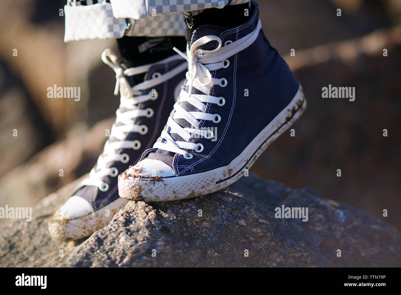 Legs, which are wearing plaid pants and blue sports dirty sneakers with white laces, standing on a rock. Stock Photo