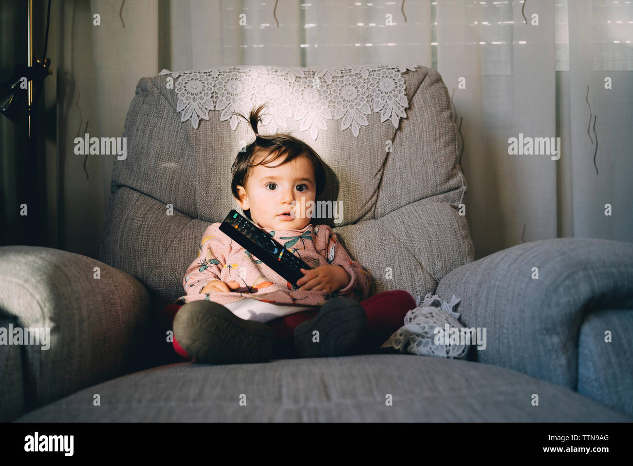 Cute baby girl with remote control sitting on armchair at home Stock Photo
