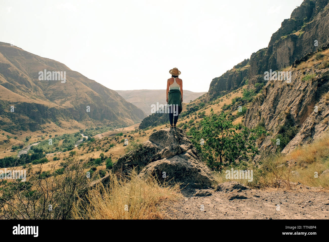 Rear view of woman looking at mountains while standing on rock against clear sky during sunny day Stock Photo