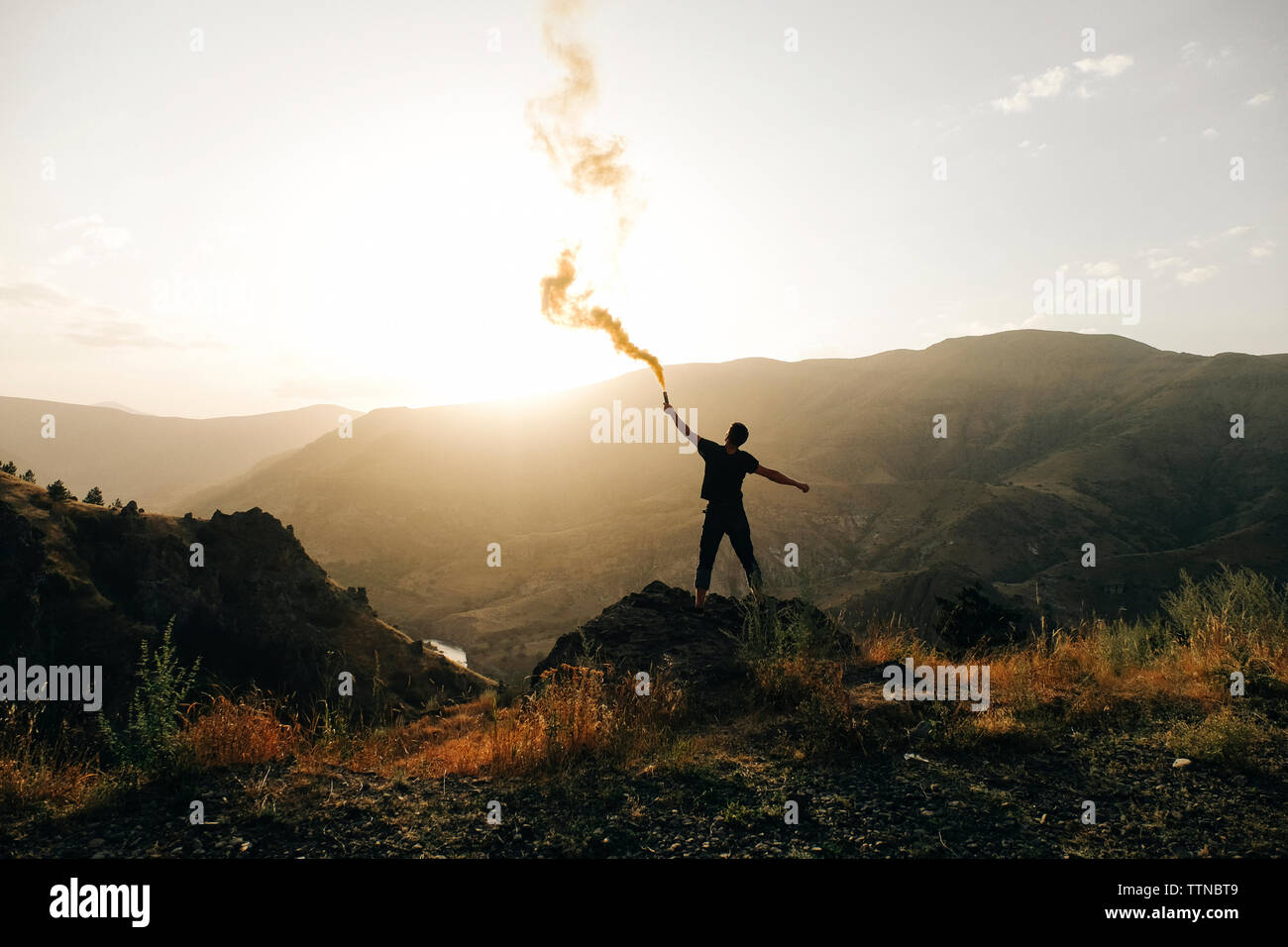 Rear view of silhouette man holding distress flare while standing on mountain against sky during sunset Stock Photo