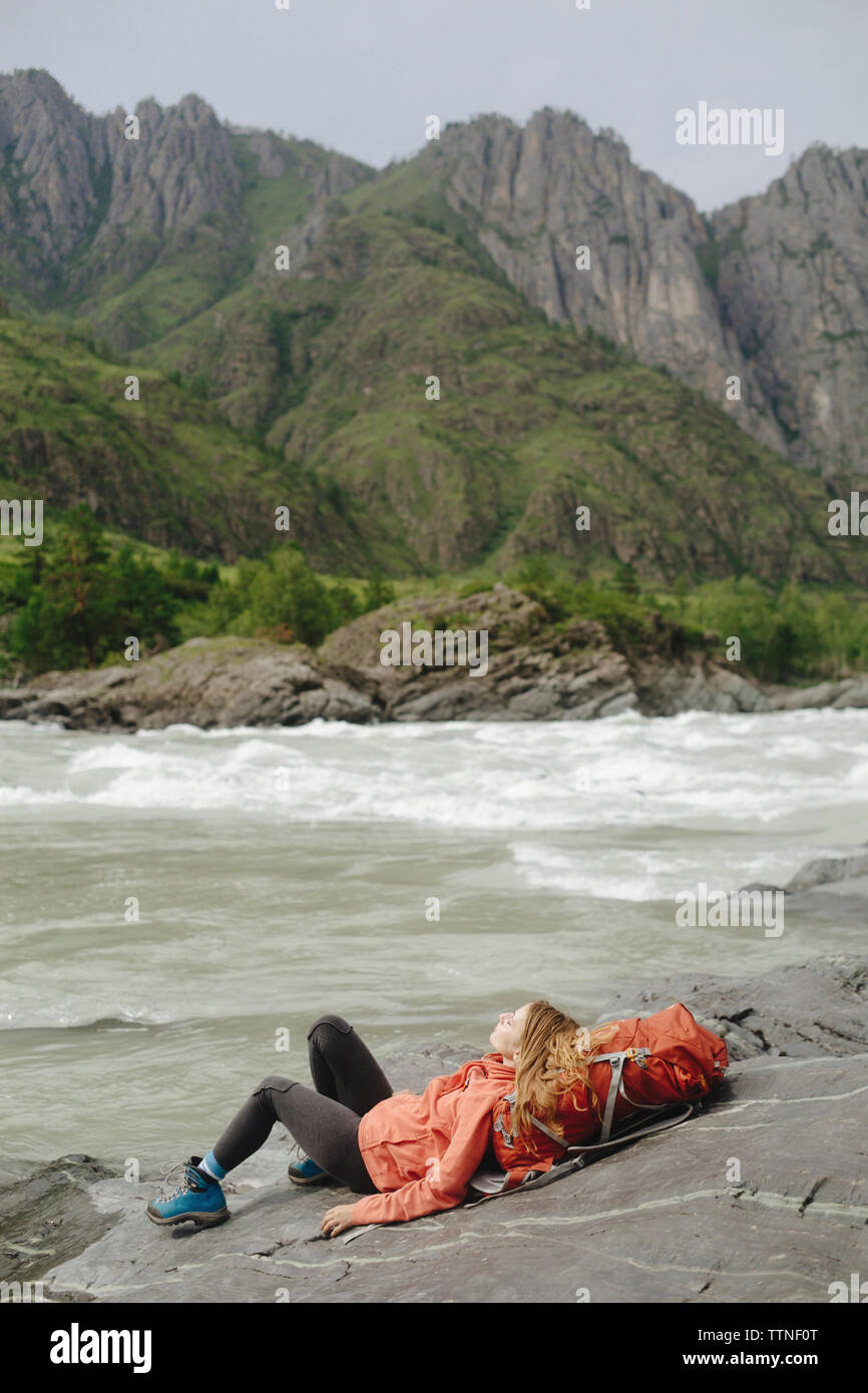 Female hiker reclining on backpack at riverbank against mountain Stock Photo