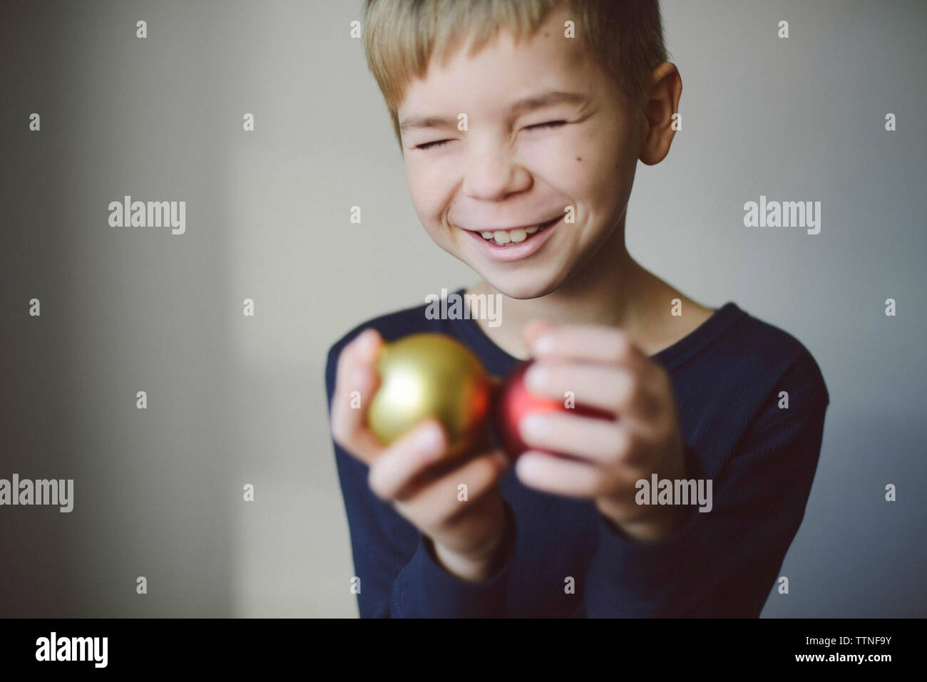 Cheerful boy with eyes closed holding christmas Baubles against wall at home Stock Photo