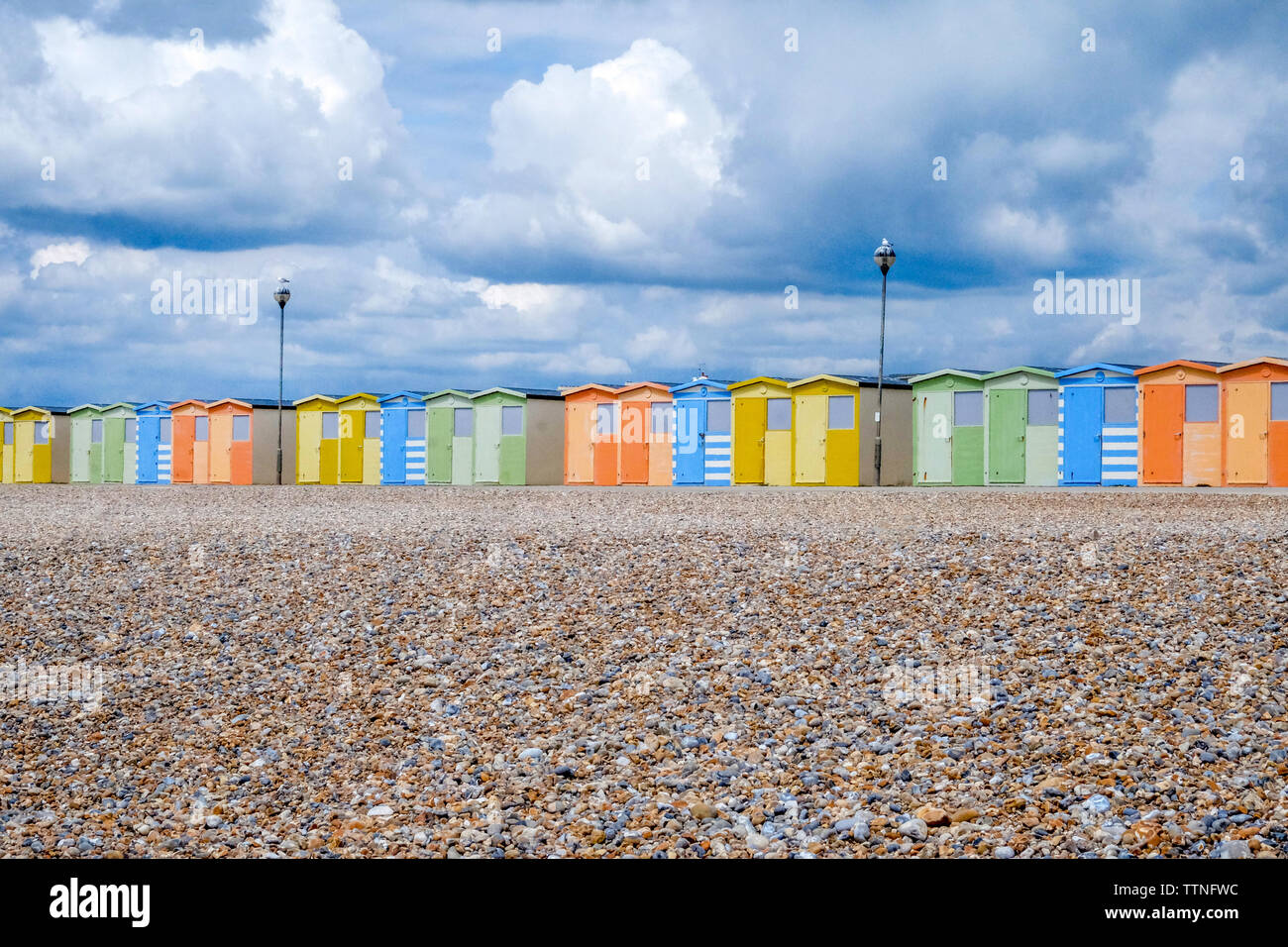 A line of 22 colourful beach huts running through the centre of the image, below is a yellow pebble beach and above is a dramatic blue and white cloud Stock Photo