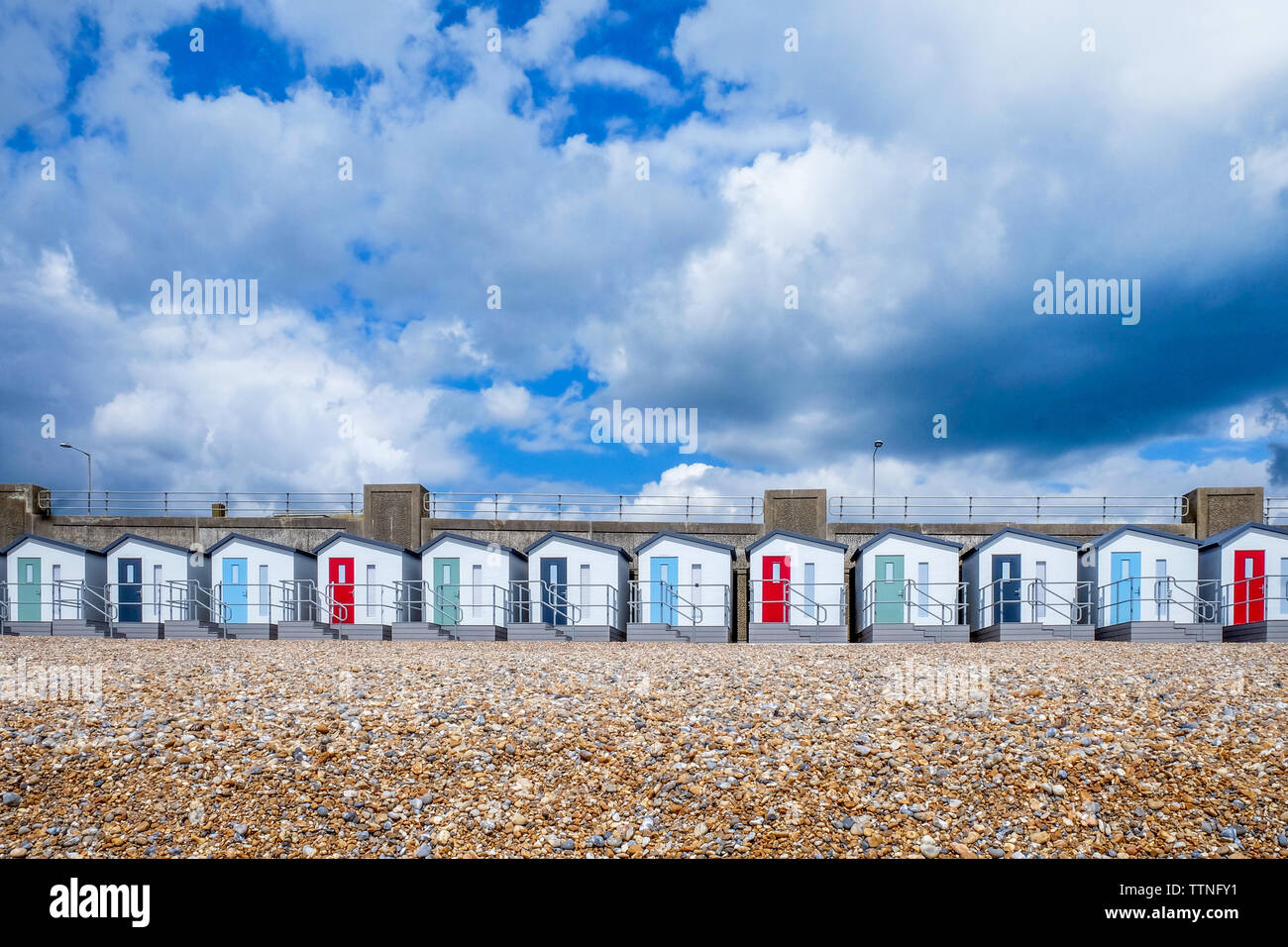 A line of 12 new Beach huts running through the centre of the image with red, blue and green doors and balconies, below is a yellow pebble beach and a Stock Photo