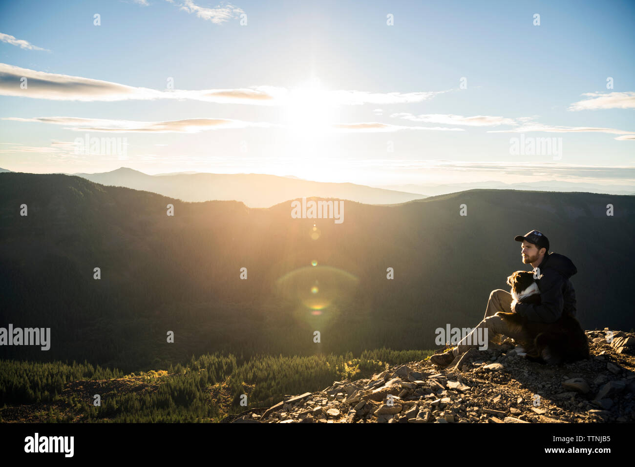 Man sitting with dog on cliff against mountain range Stock Photo