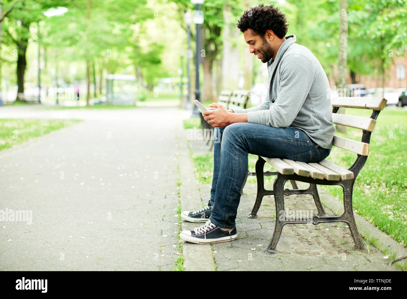 Side view of happy man using smart phone while sitting on park bench by  footpath Stock Photo - Alamy