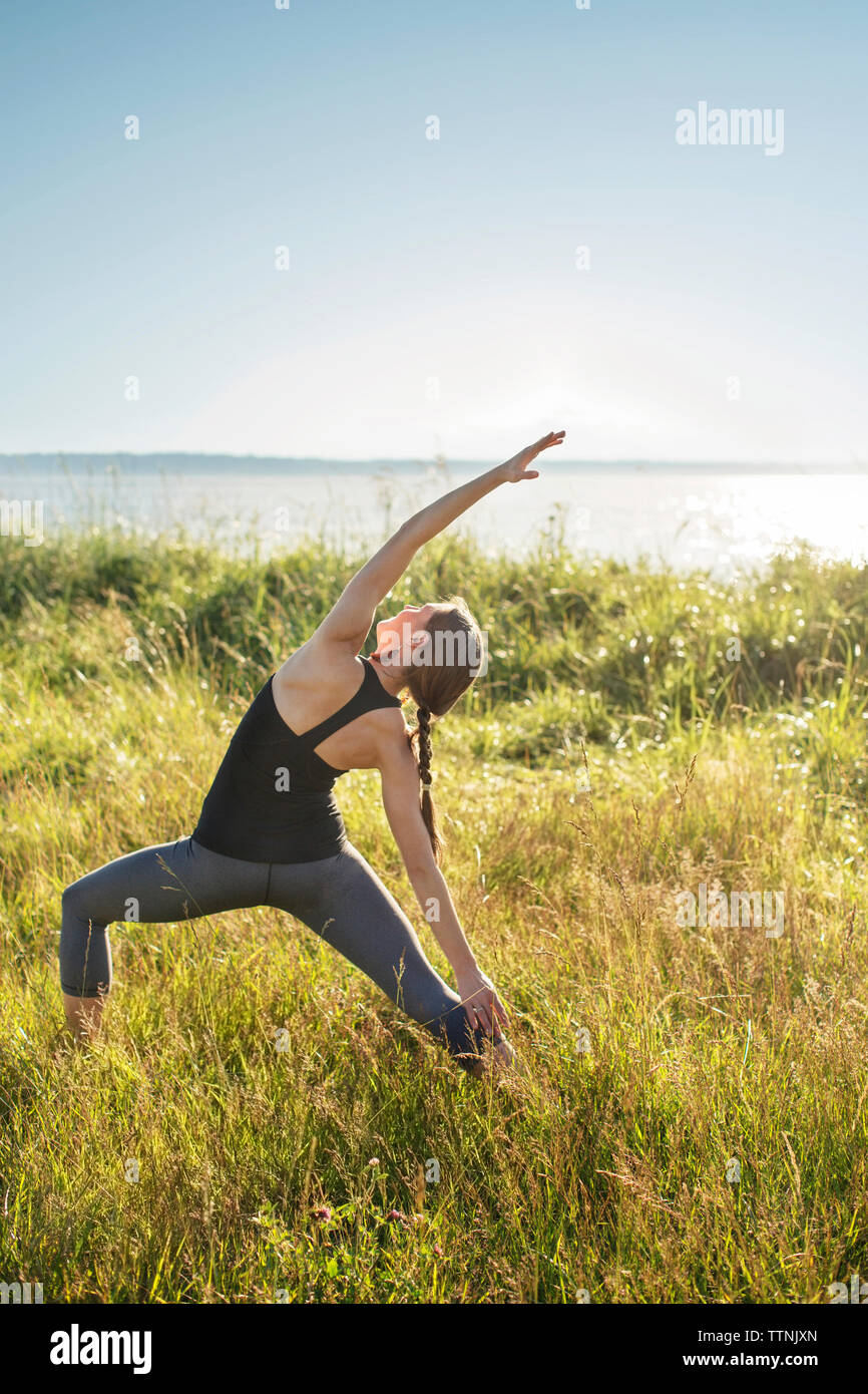 Woman practicing extended side angle pose yoga on grassy field by sea during sunny day Stock Photo