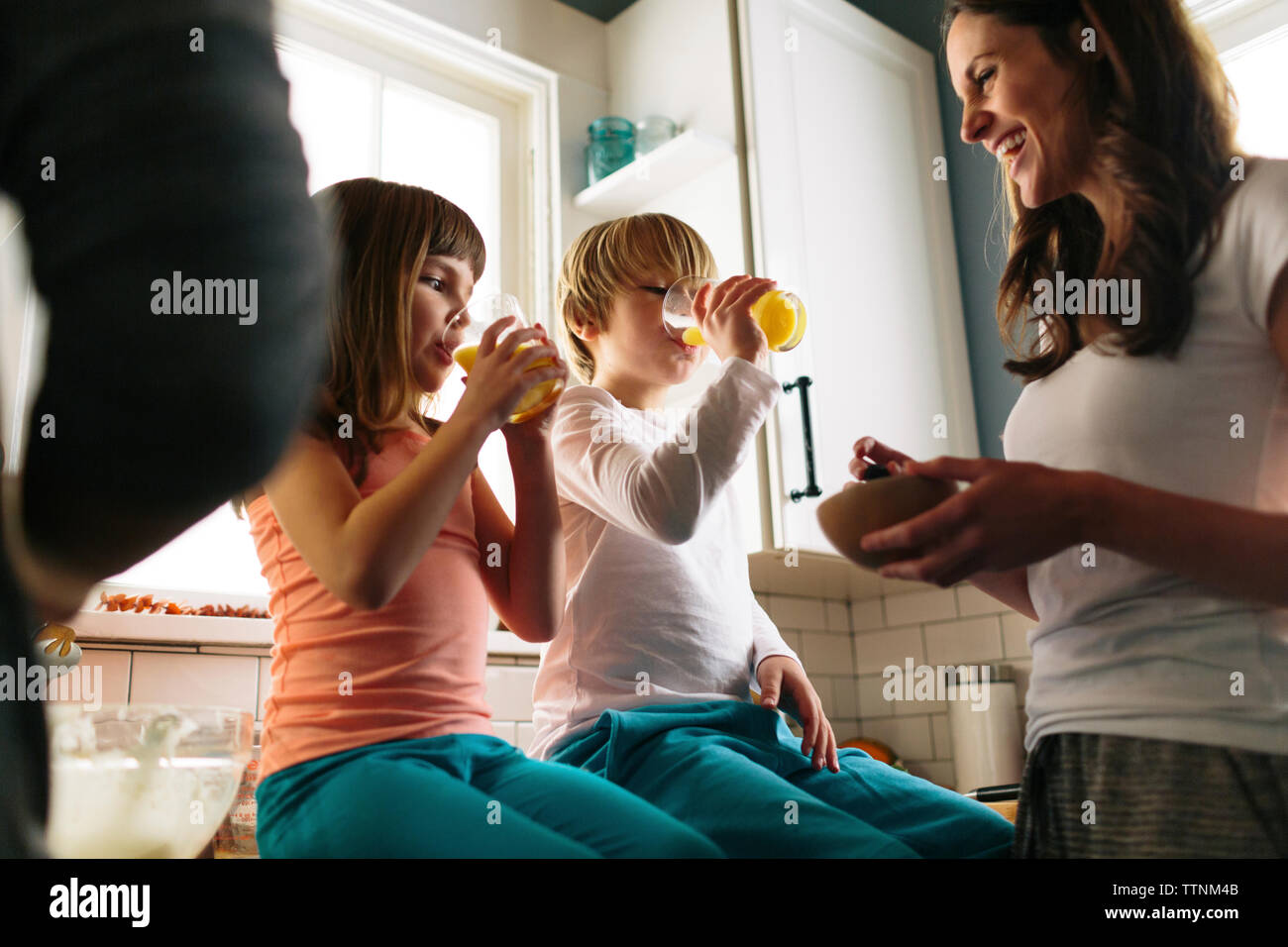 Mother looking at children drinking juice while sitting on kitchen counter Stock Photo