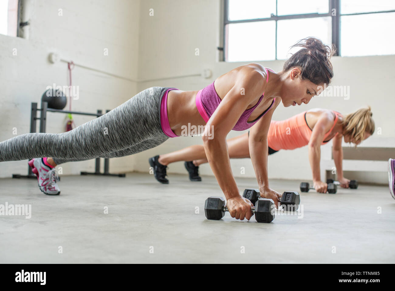 Female athletes holding dumbbells while doing push-ups in gym Stock Photo